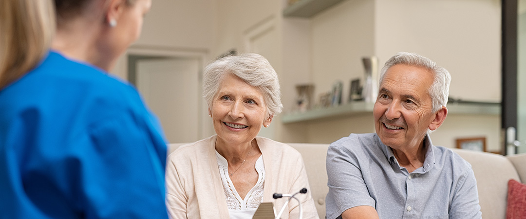 Senior couple sitting on sofa talking to healthcare worker in blue scrubs.